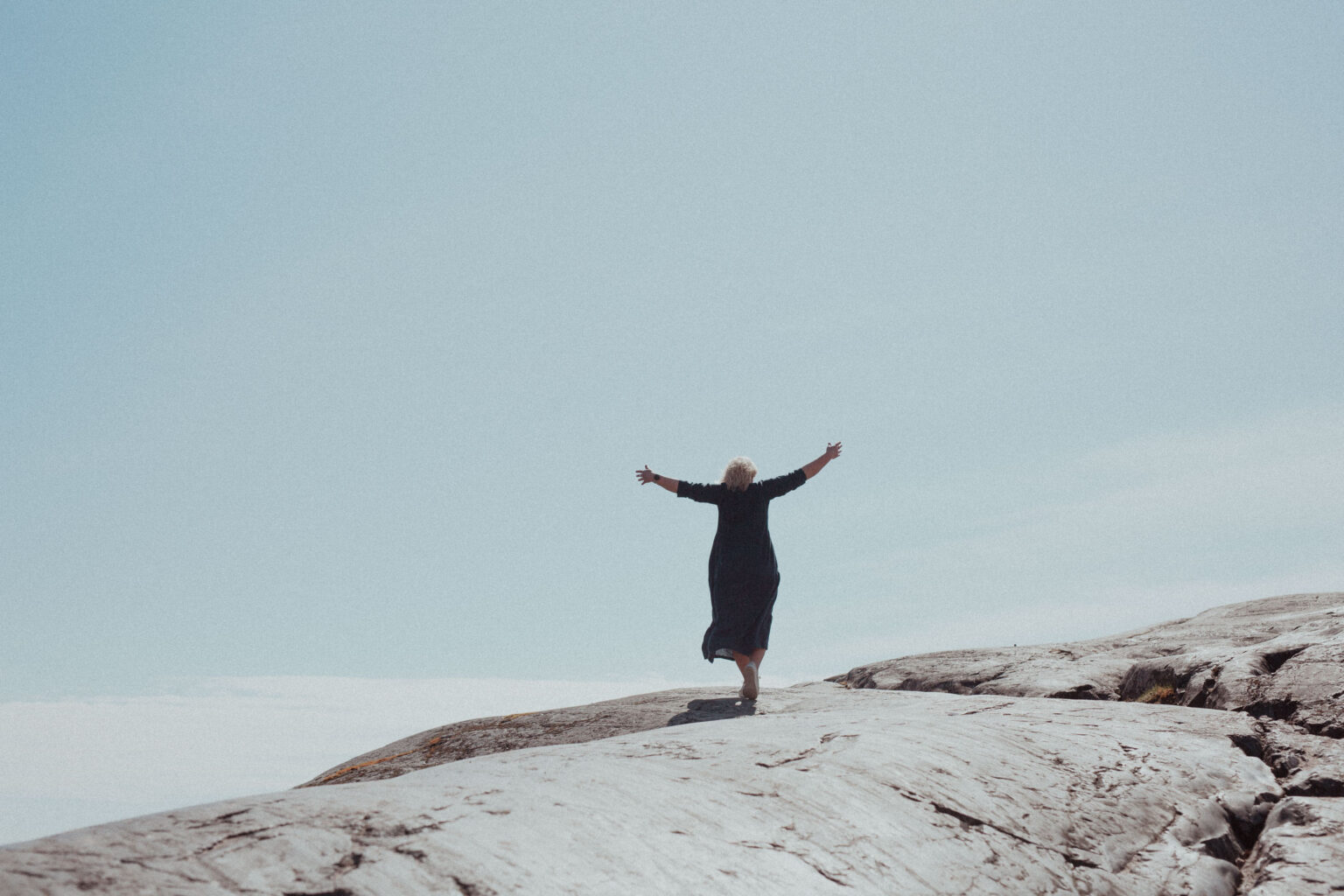 Nainen seisoo kalliolla molemmat kädet vapautuneesti levitettynä sivuille katsellen samalla levollista näkymää merelle ja nauttii jälleen elämästään, minkä mahdollisti terapiapalvelut.