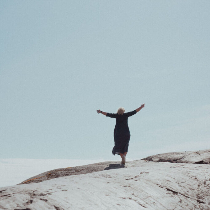 Nainen seisoo kalliolla molemmat kädet vapautuneesti levitettynä sivuille katsellen samalla levollista näkymää merelle ja nauttii jälleen elämästään, minkä mahdollisti terapiapalvelut.