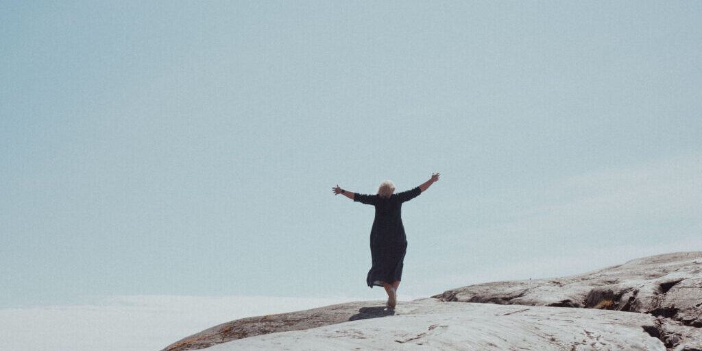 Nainen seisoo kalliolla molemmat kädet vapautuneesti levitettynä sivuille katsellen samalla levollista näkymää merelle ja nauttii jälleen elämästään, minkä mahdollisti terapiapalvelut.